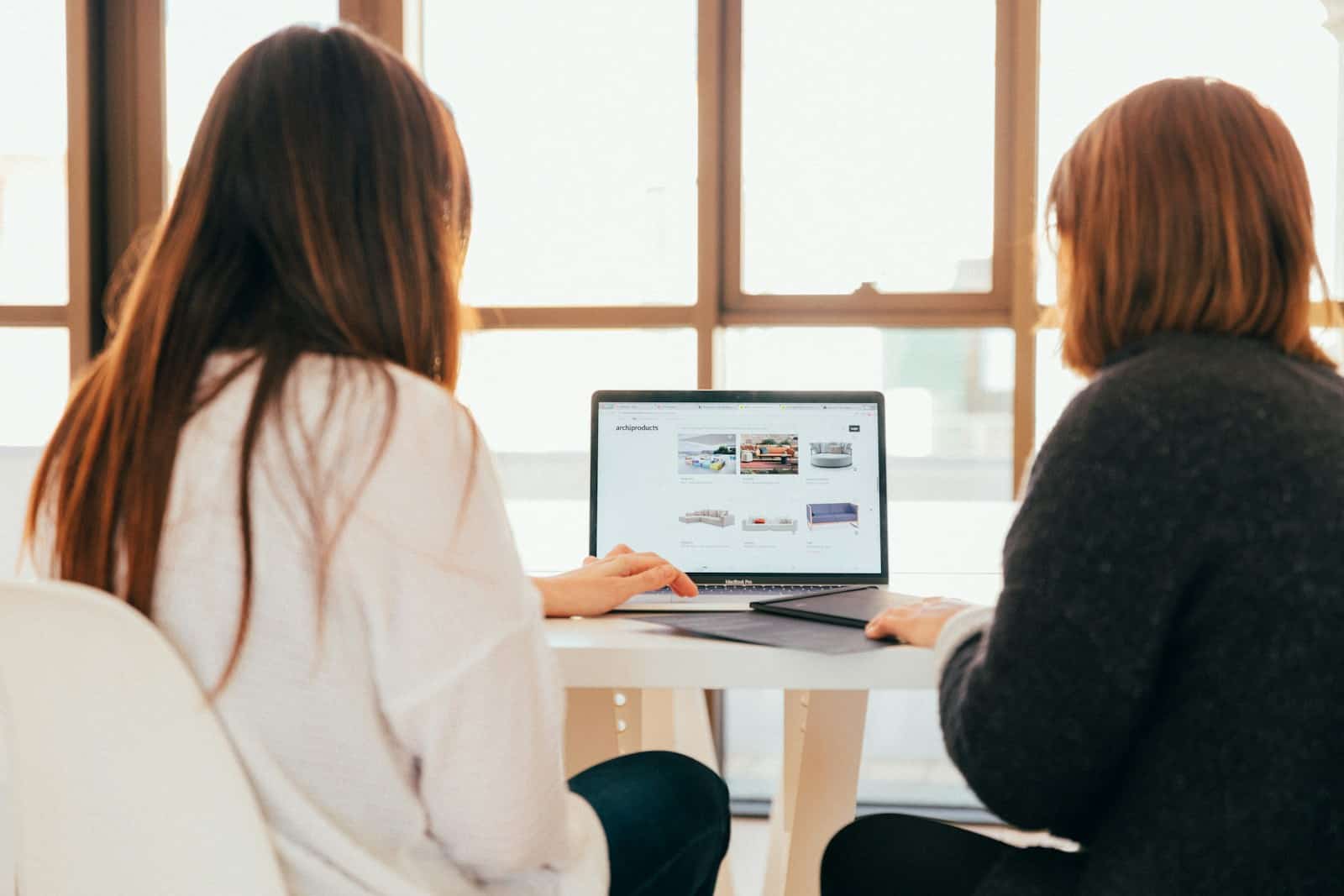 Two people sit at a table browsing furniture on a laptop. Large windows are in the background.
