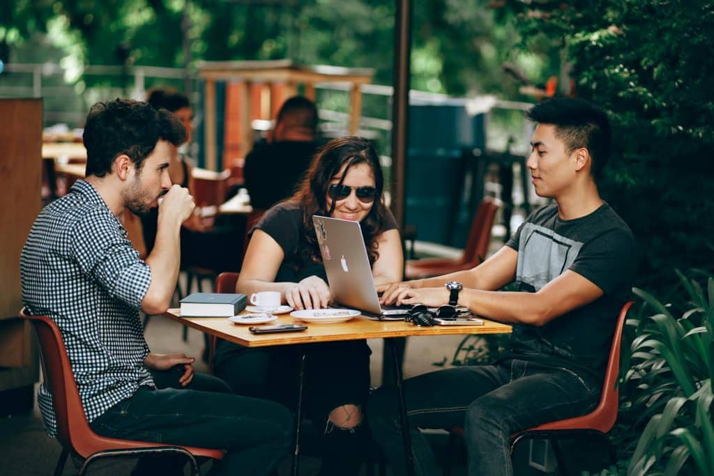 Three people sitting outdoors at a café, using a laptop, with coffee cups on the table.