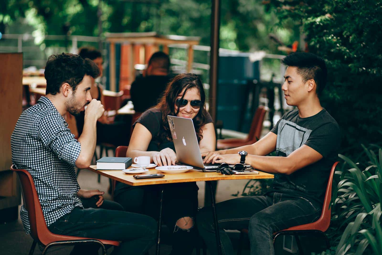 Three people sitting outdoors at a café, using a laptop, with coffee cups on the table.