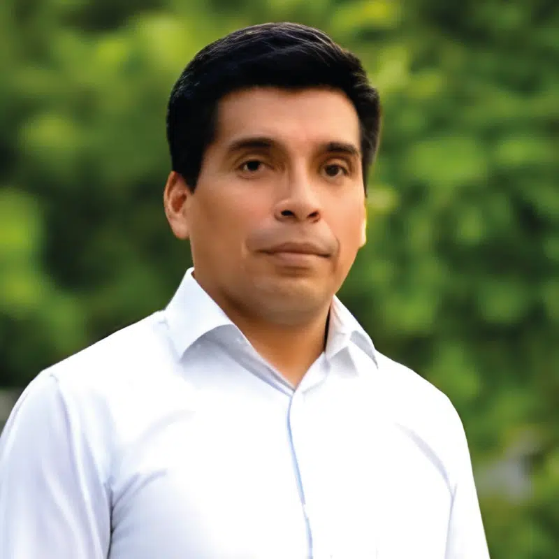 Man in a white shirt stands outdoors against a blurred green background.