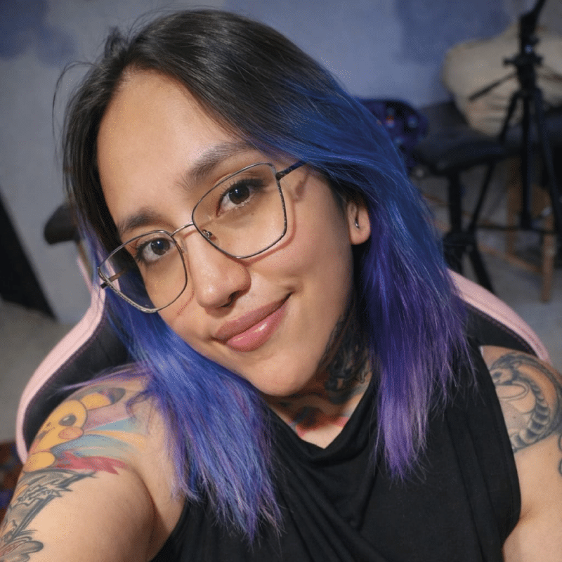 Person with blue and purple hair, glasses, tattoos, and a black top smiles at the camera.