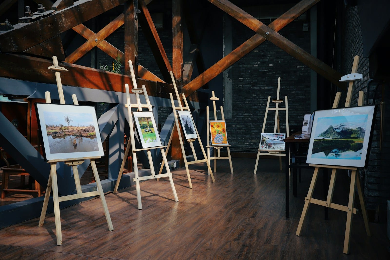Art exhibition with framed paintings on wooden easels in a dimly lit gallery space.