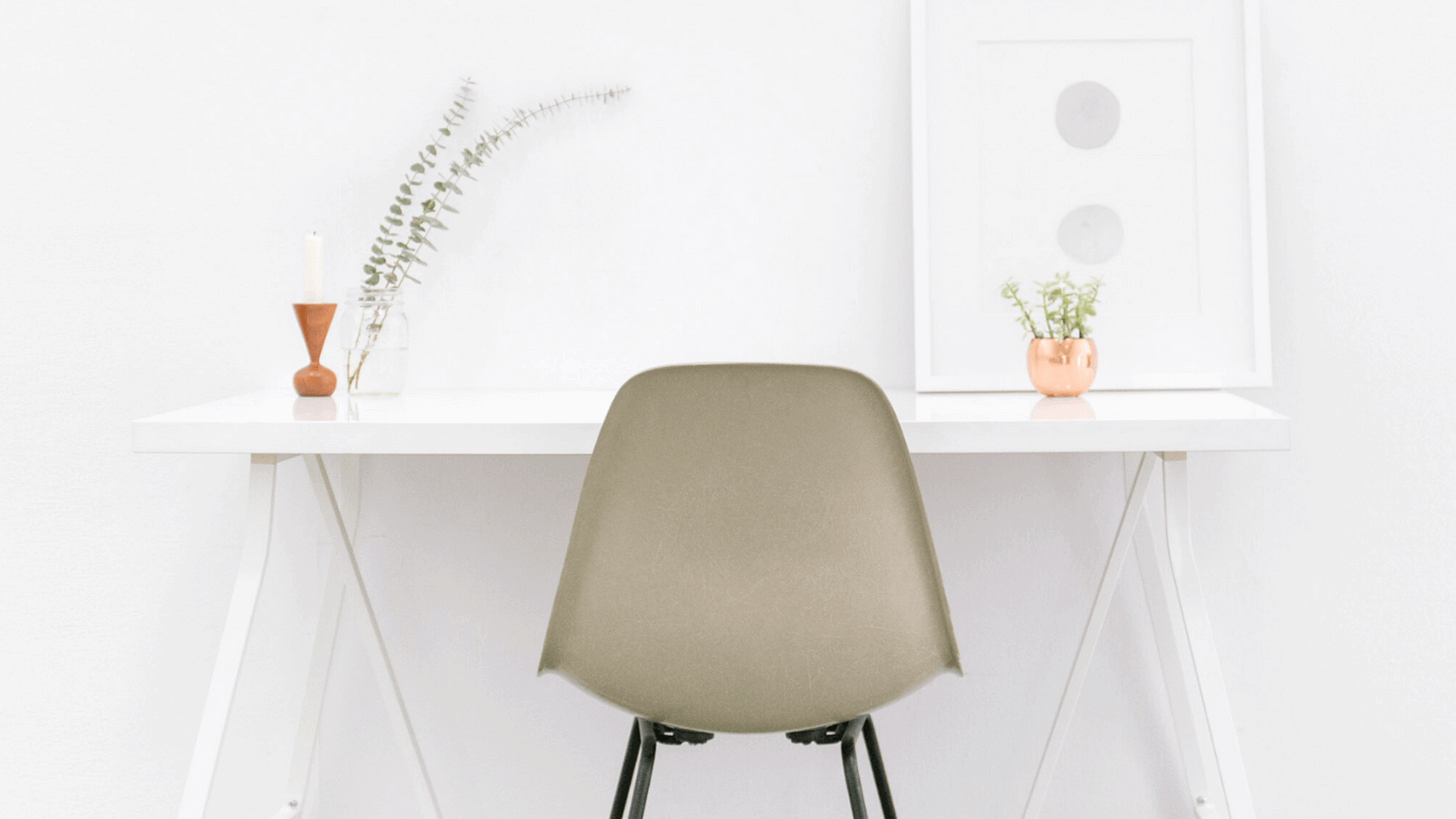Minimalist desk with a gray chair, greenery, candle, and art frame against a white wall.