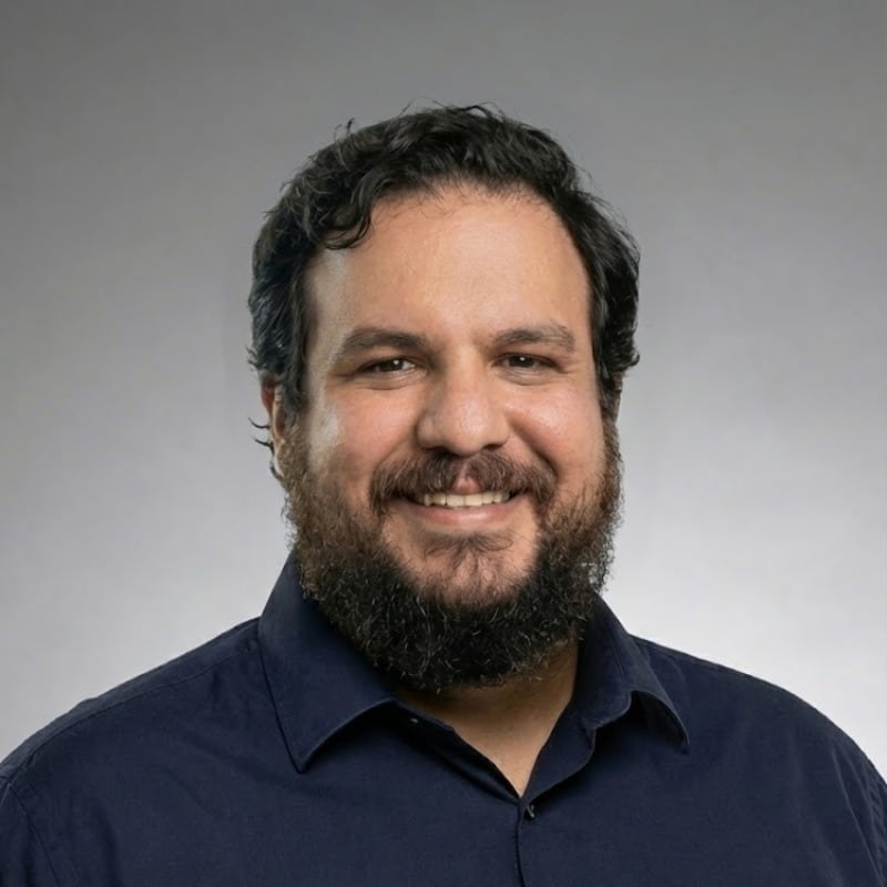 Smiling man with a beard and dark hair, wearing a dark shirt, on a gray background.