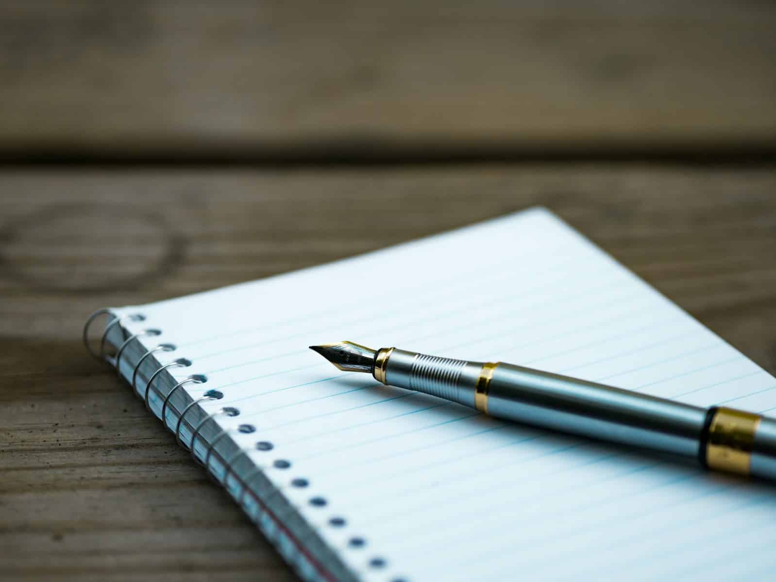 A fountain pen rests on an open spiral notebook with lined paper, on a wooden surface.