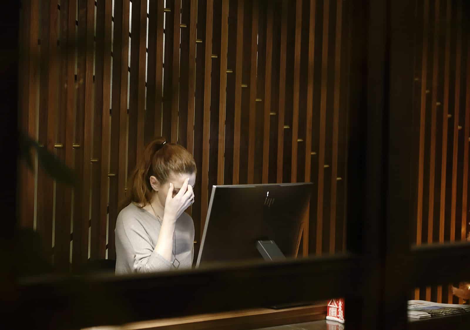 Person with hand on face, sitting at a desk with a computer, in a room with wooden paneling.