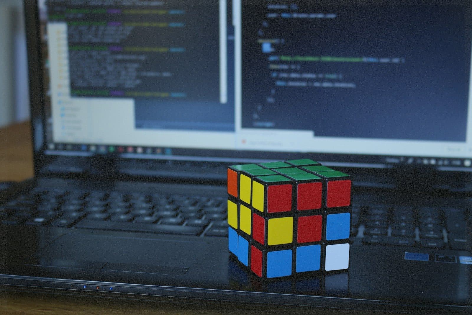 A Rubik's Cube on a laptop keyboard with code on the screen in the background.